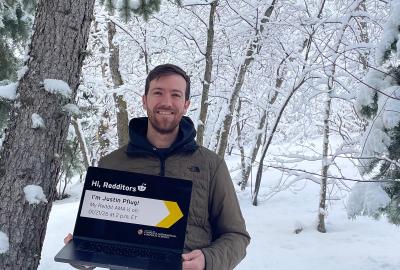 Justin Pflug wearing a coat and holding a laptop in front of a snowy forest