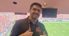 Cole Miller giving two thumbs up standing in stadium with a football field in the background