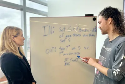 A woman, Dr. Erin Molloy, and a man (her student) are observing a whiteboard.