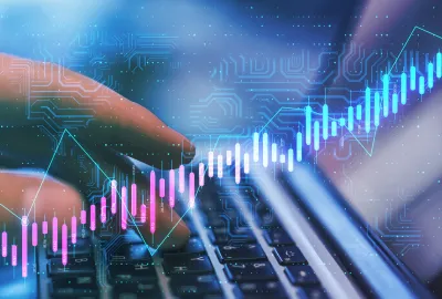 A close-up view of a person's hands typing on a laptop keyboard, overlaid with a glowing digital graphic of a rising financial stock chart, data points, and circuit board patterns in shades of blue, pink, and turquoise.