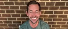 A man with brown hair and facial hair smiles wearing a dark green shirt in front of a brick wall. 