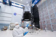 Jackie Townsend wearing all-white lab gear and blue gloves, gesturing to the Roman Space Telescope behind her