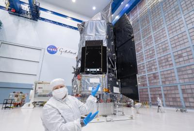 Jackie Townsend wearing all-white lab gear and blue gloves, gesturing to the Roman Space Telescope behind her