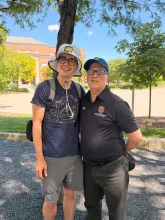 Two men stand outside, smiling on a sunny day. The one on the left wears gray shorts, a navy t-shirt, and a light gray bucket hat. The one on the right wears black jeans, a black University of Maryland promo, and a blue baseball cap.
