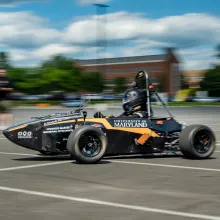 A black racecar with the text "University of Maryland" moves along a blurry background of a blue, cloudy sky and green trees.