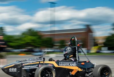 A black racecar with the text "University of Maryland" moves along a blurry background of a blue, cloudy sky and green trees.