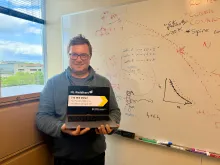 A man holding a laptop in front of a window and a whiteboard