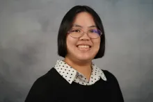 A young woman in a black-and-white polka dot button-down shirt and a black sweater smiles in front of a mottled gray backdrop.