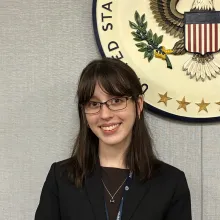 A young woman wearing a dark blazer and NRC lanyard, smiles in front of the U.S. Nuclear Regulatory Commission seal, with an American flag beside her.