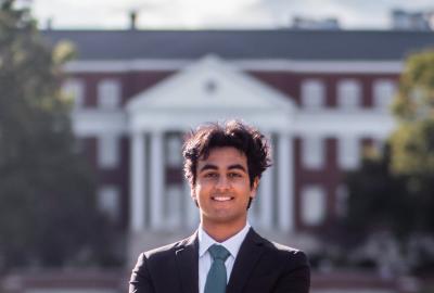 Dhruv Dewan stands in a suit in front of a red academic building.