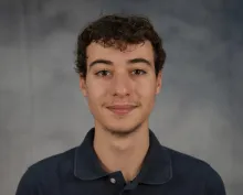 Headshot of a man with curly hair wearing a navy blue polo shirt in front of a mottled gray backdrop. 