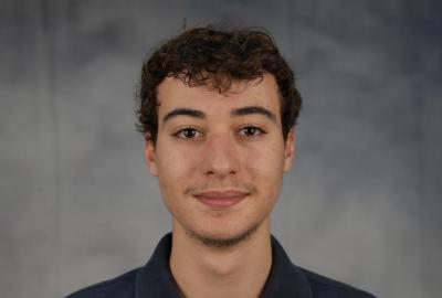 Headshot of a man with curly hair wearing a navy blue polo shirt in front of a mottled gray backdrop. 
