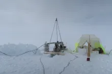 A snowy terrain. A metal drilling device rests on plywood over a hole in snow. Next to it is a white and neon yellow tent housing scientific equipment.