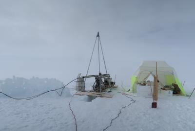 A snowy terrain. A metal drilling device rests on plywood over a hole in snow. Next to it is a white and neon yellow tent housing scientific equipment.