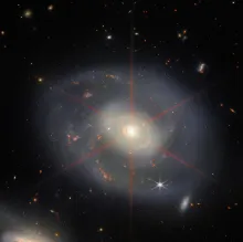 JWST image of spiral galaxy NGC 7469, featuring a white core that emits red six-pointed lines across its center. Faint-blue spiral arms dotted with orange-red dots sit against the dark background of space.