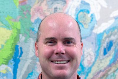 A man in a plaid shirt stands in front of a colorful map.