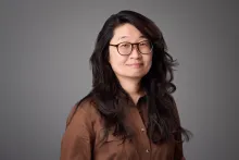 Portrait of a woman wearing glasses and a brown button-down shirt against a neutral grey background.