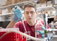 A man in a red t-shirt and glasses wears lab gloves and pipettes liquids. 