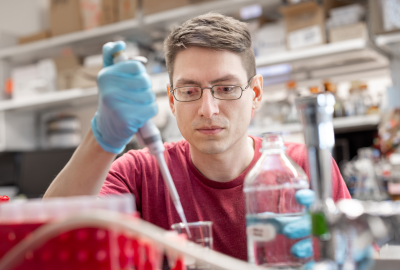 A man in a red t-shirt and glasses wears lab gloves and pipettes liquids. 