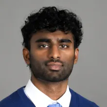 A headshot of a man in a white dress shirt, baby blue tie and royal blue sweater. He is situated in front of a gray background.