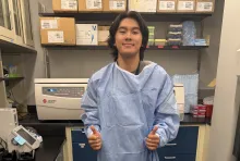 A young man wearing an oversized blue medical gown smiles and gives two thumbs-up inside a laboratory filled with equipment, supply boxes, and shelving.