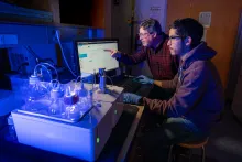 Two researchers work on a computer in a dimply lit room. One older man wears a checkered red button-down shirt, while a younger man wears a maroon hoodie. Both wear glasses. In front of them are beakers and flasks illuminated in blue light.
