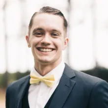 A man smiles wearing a black suit and yellow bow tie.