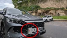 Close-up of a dark grey BYD vehicle parked on a road next to an old stone wall structure. Encircled in red on the front bumper is a small grey plastic box with a red "2" written on it, secured to the front grill next to the UK license plate.