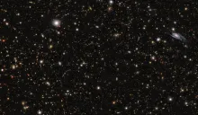 Telescope image depicting a black sky with hundreds of red, yellow, white and blue dots, plus a blue galaxy in the top-right of the image.