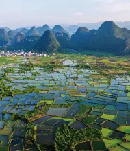 Aerial view of a patchwork of flooded rice paddies in a valley in China. In the background is a small village of white and orange buildings in front of towering karst peaks. All of this sits below a hazy sky.