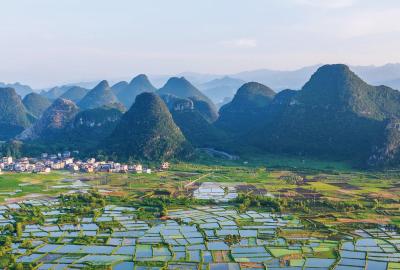 Aerial view of a patchwork of flooded rice paddies in a valley in China. In the background is a small village of white and orange buildings in front of towering karst peaks. All of this sits below a hazy sky.