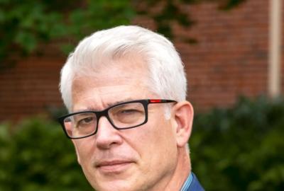 A man in black glasses and a blue suit over a blue plaid shirt stands in front of blurred greenery and bricks.