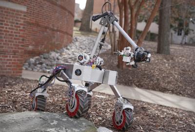 A small four-wheeled robotic rover with a mounted mechanical arm drives over rocks outdoors, near a brick building and leafless trees.