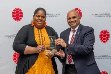 A woman in a black and yellow dress accepts a glass award from a man in a blue suit with a white shirt and red tie. They stand in front of a backdrop with red turtle shells and text saying "College of Computer, Mathematical, & Natural Sciences."