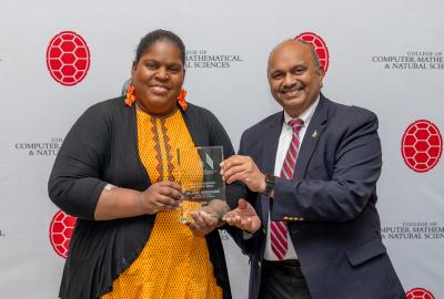 A woman in a black and yellow dress accepts a glass award from a man in a blue suit with a white shirt and red tie. They stand in front of a backdrop with red turtle shells and text saying "College of Computer, Mathematical, & Natural Sciences."