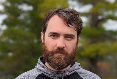 A bearded, brunette man wears a black and gray hoodie that says "SEED Electronic Arts" standing in front of a blurred background of trees.