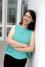 A young woman leans against a white column outdoors, smiling with her arms crossed. She wears a blue, sleeveless shirt and black pants. Glass doors are blurred in the background.