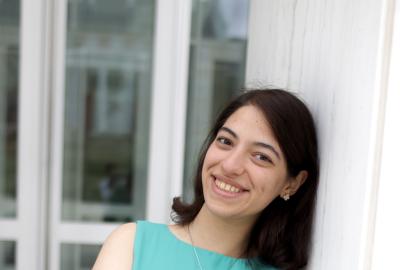 A young woman leans against a white column outdoors, smiling with her arms crossed. She wears a blue, sleeveless shirt and black pants. Glass doors are blurred in the background.