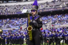A member of a marching band stands on a field holding a tenor saxophone, wearing a detailed purple and black uniform with gold trim, a black hat topped with a tall purple feather plume, and dark sunglasses. The blurred background shows a crowded stadium seating area under a "30 SEASONS" sign and other band members lined up on the field.