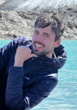 Brown haired man with beard and blue jacket smiling with his chin on his hand in front of water and rocks.