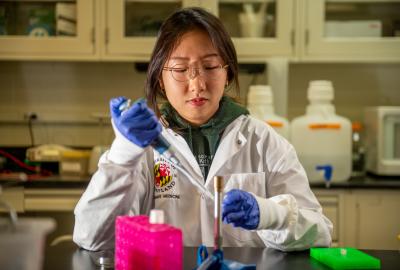 A woman in a lab coat wearing blue gloves and glasses uses a pipette to transfer liquic into a test tube.