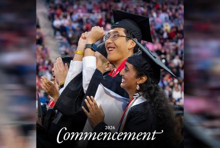 Two students in graduation regalia celebrating, with a blurred crowd in the background.