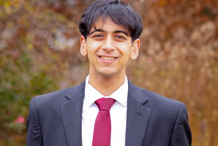 A male college-aged student wearing a black suit with a white dress shirt and red tie stands in front of a blurred background of autumnal-colored foliage.