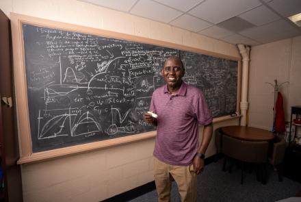 Professor Abba Gumel. wearing a red polo and khaki pants, holds a piece of chalk smiling in front of a chalkboard filled with mathematics equations and graphs. 