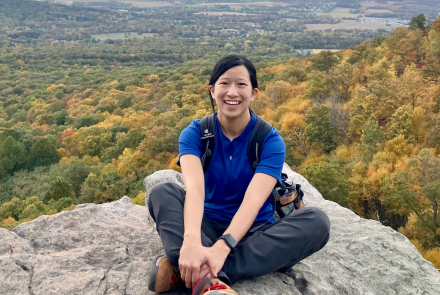 Biological sciences graduate student Stephanie Chia sits smiling at the camera on a rock overlooking a cloudy sky and a valley of trees.