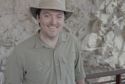 Nathan Jud in brown hat and shirt smiles while standing in front of a rock wall.
