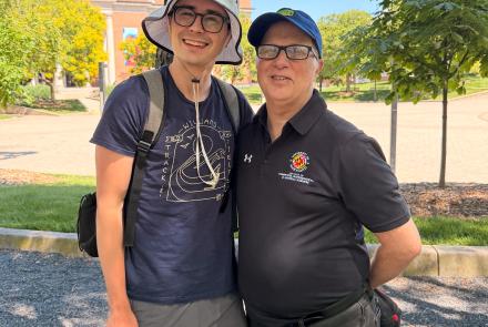 Two men stand outside, smiling on a sunny day. The one on the left wears gray shorts, a navy t-shirt, and a light gray bucket hat. The one on the right wears black jeans, a black University of Maryland promo, and a blue baseball cap.