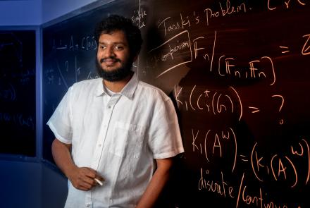 Srivatsav Kunnawalkam Elayavalli holding a piece of chalk and standing in front of a blackboard full of equations