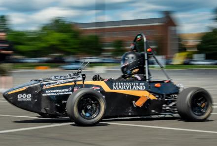 A black racecar with the text "University of Maryland" moves along a blurry background of a blue, cloudy sky and green trees.