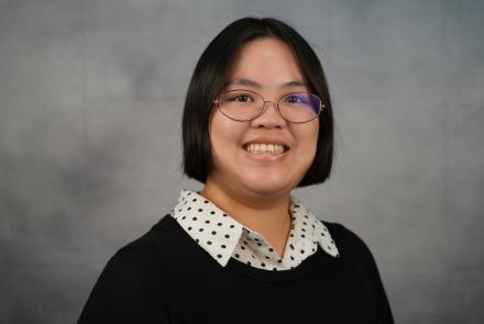 A young woman in a black-and-white polka dot button-down shirt and a black sweater smiles in front of a mottled gray backdrop.